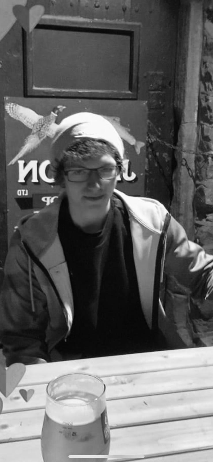 Person sitting at a table with a drink, wearing glasses and a hoodie, in front of a branded backdrop.